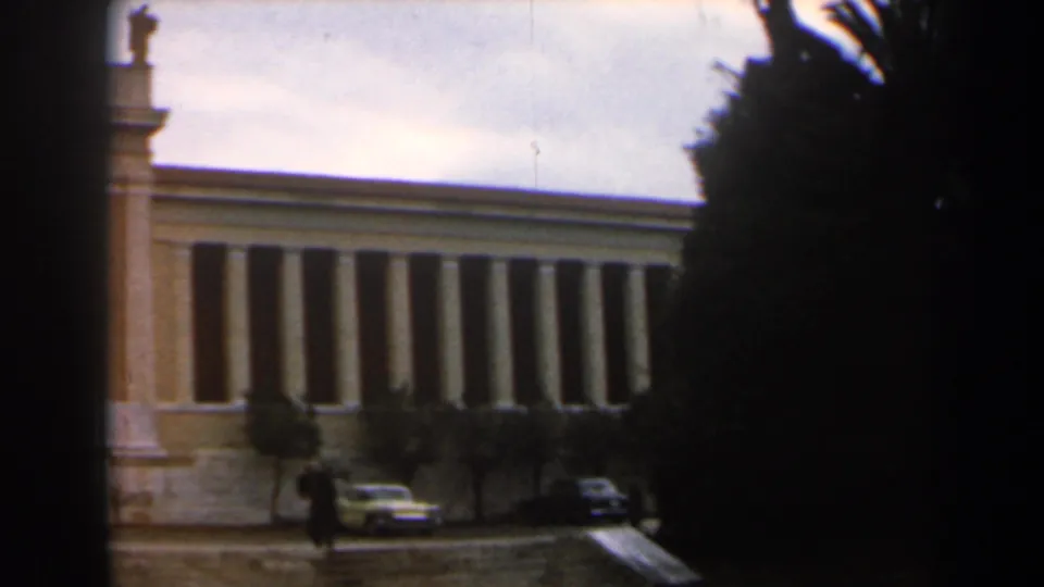 1960 Israel 8mm frame showing a colonnaded civic-looking exterior with a statue, parked cars, trees, and distant pedestrians.