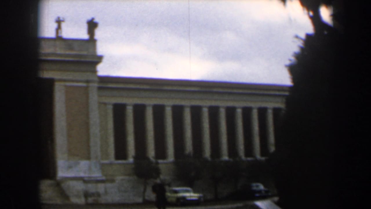 Tan colonnaded building under cloudy sky with cars and a distant pedestrian in front.