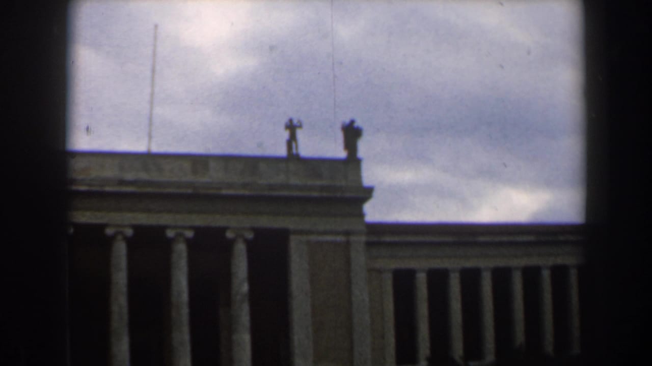 Upper facade of a tan building with columns and silhouetted rooftop statues.