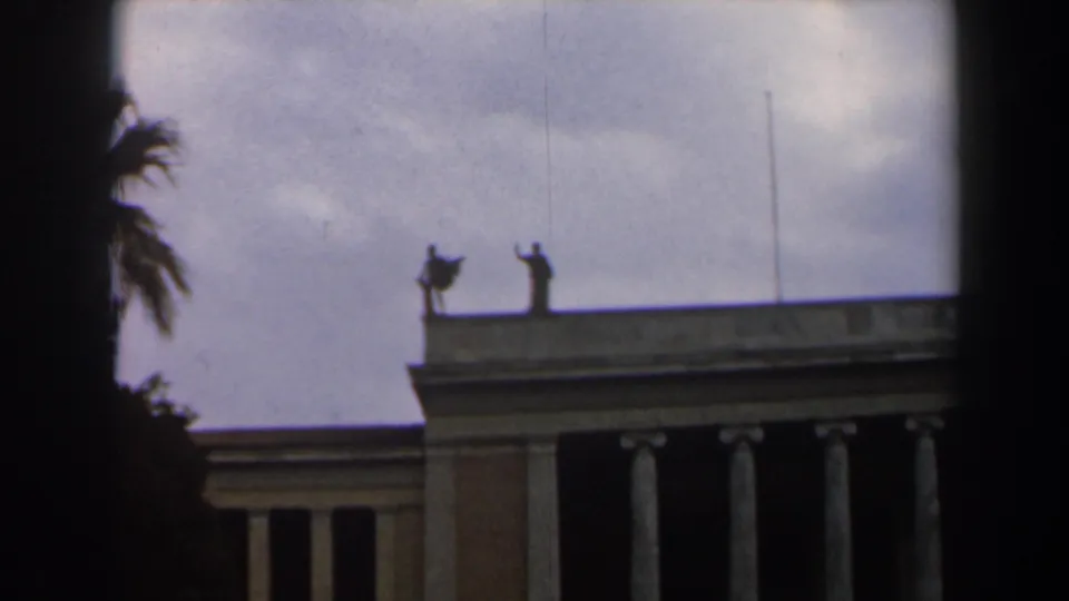 1960 Israel 8mm frame showing rooftop statues above a tan colonnaded building, with palm fronds and clouds nearby.