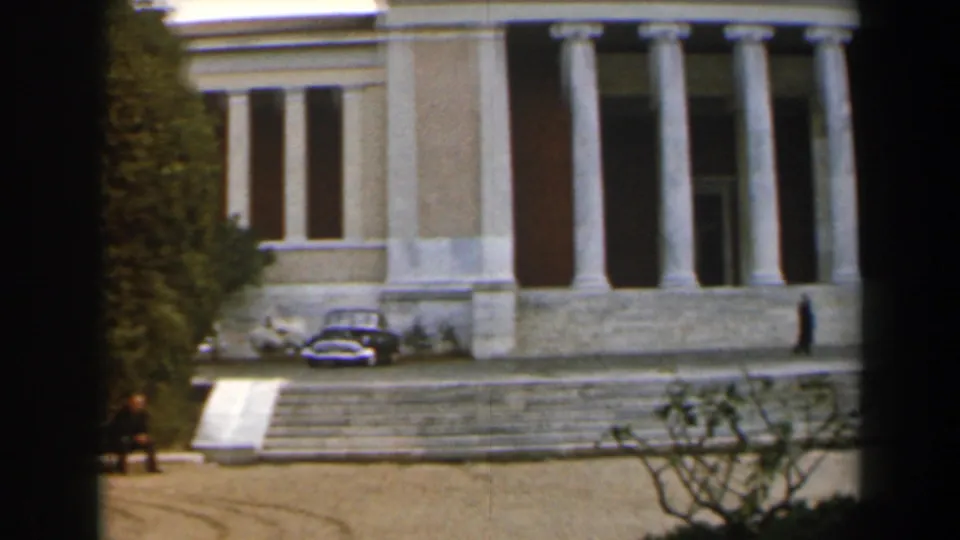 1960 Israel home-movie frame showing the columned entrance, stone steps, parked cars, and small pedestrians near a civic building.