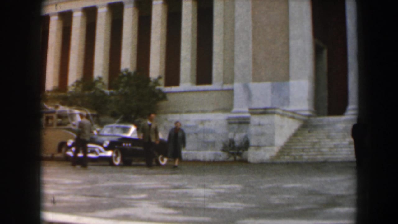 Adults near parked cars outside a large columned building.