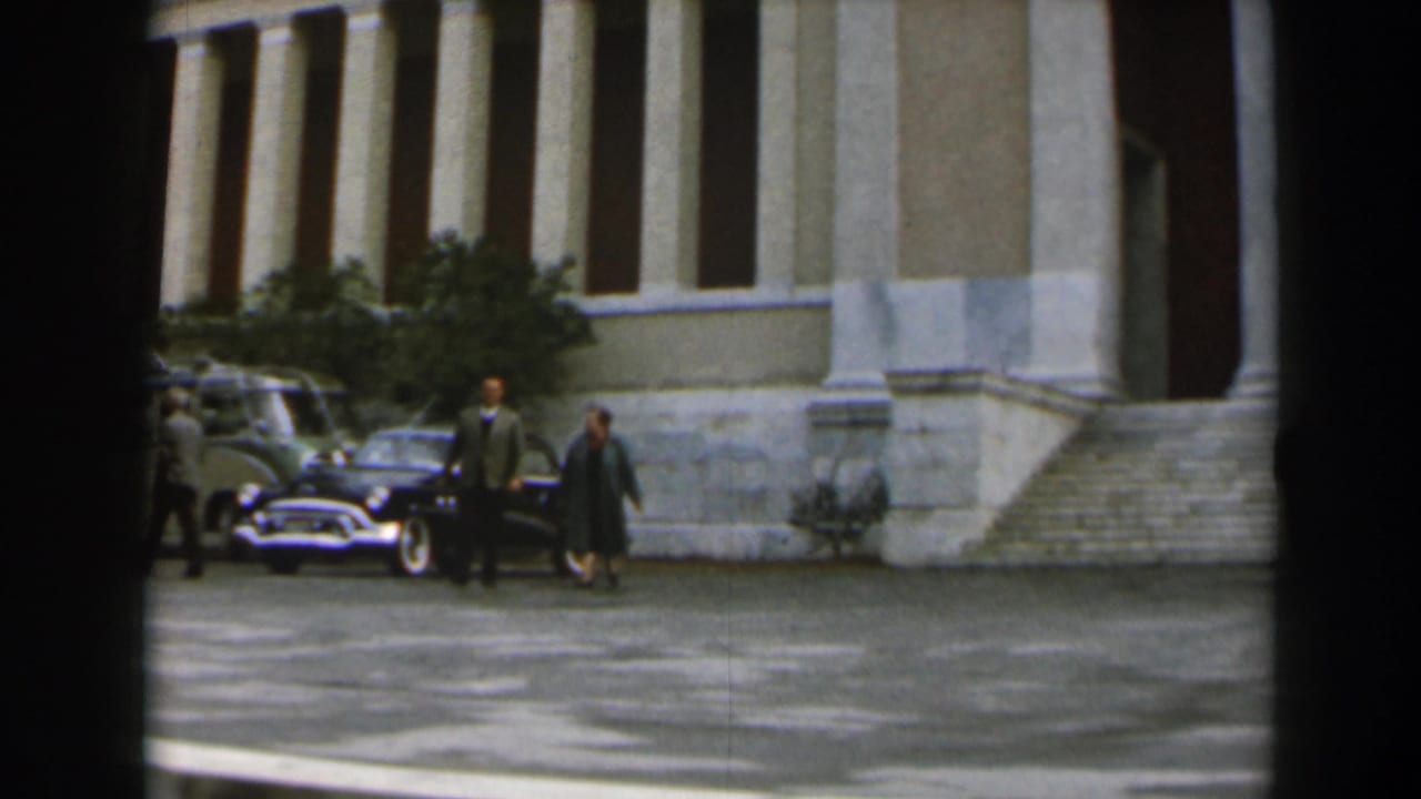 Two adults walk away from parked cars near a columned entrance.