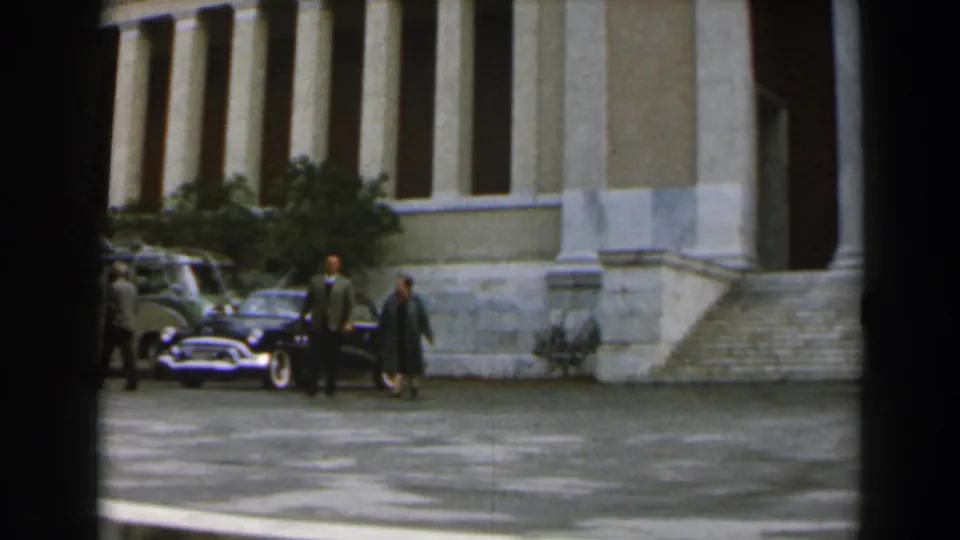 Two adults in formal outerwear walk across a paved courtyard near parked cars in 1960 Israel footage.