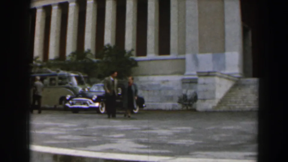 Adults walk past a dark car and light station wagon outside a tall-columned building in Israel home-movie footage.