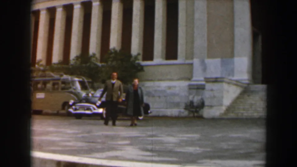Two adults walk toward the camera across a paved courtyard, with period cars and a columned building behind them.