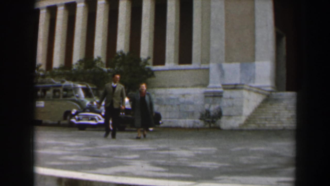 Man and woman walk across a paved courtyard in front of a columned building and parked cars.