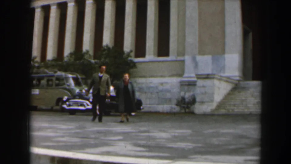 In 1960s Israel home-movie footage, two adults walk near a classical columned building with mid-century cars behind them.