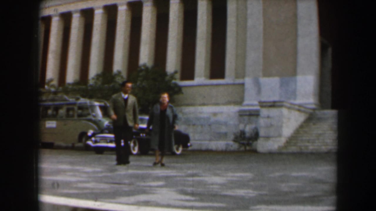 Two adults continue walking toward the camera with a station wagon and dark car behind them.
