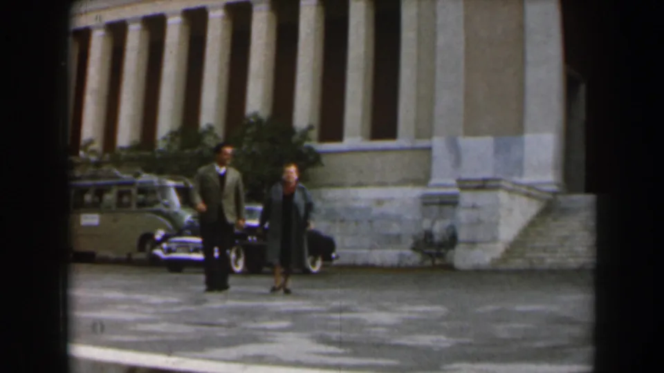 The man and woman move side by side in front of a formal columned building, with vintage parked vehicles in the background.