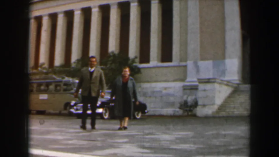 Closer view of two adults walking together in a paved courtyard outside a large columned building in Israel.