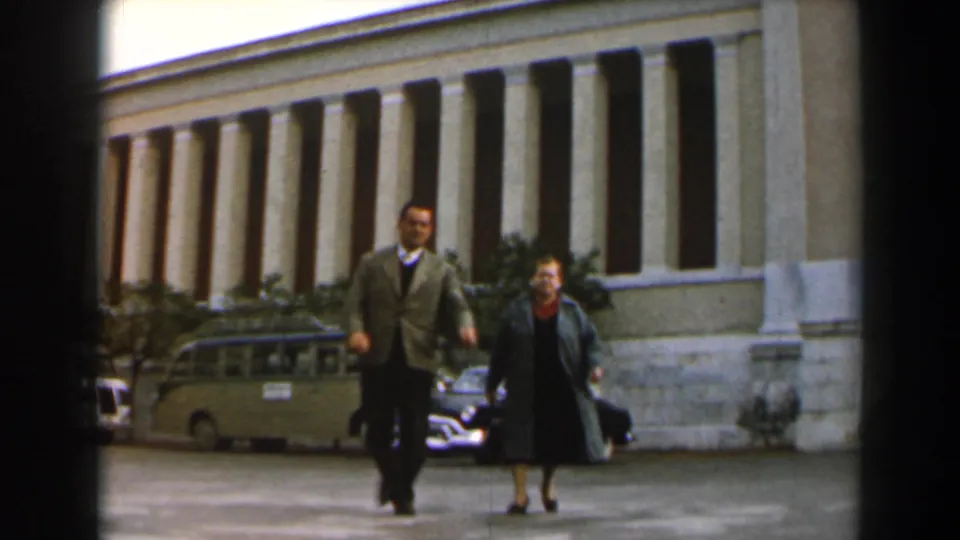 Near the end of the shot, the pair passes close to camera against the building facade and parked vehicles.