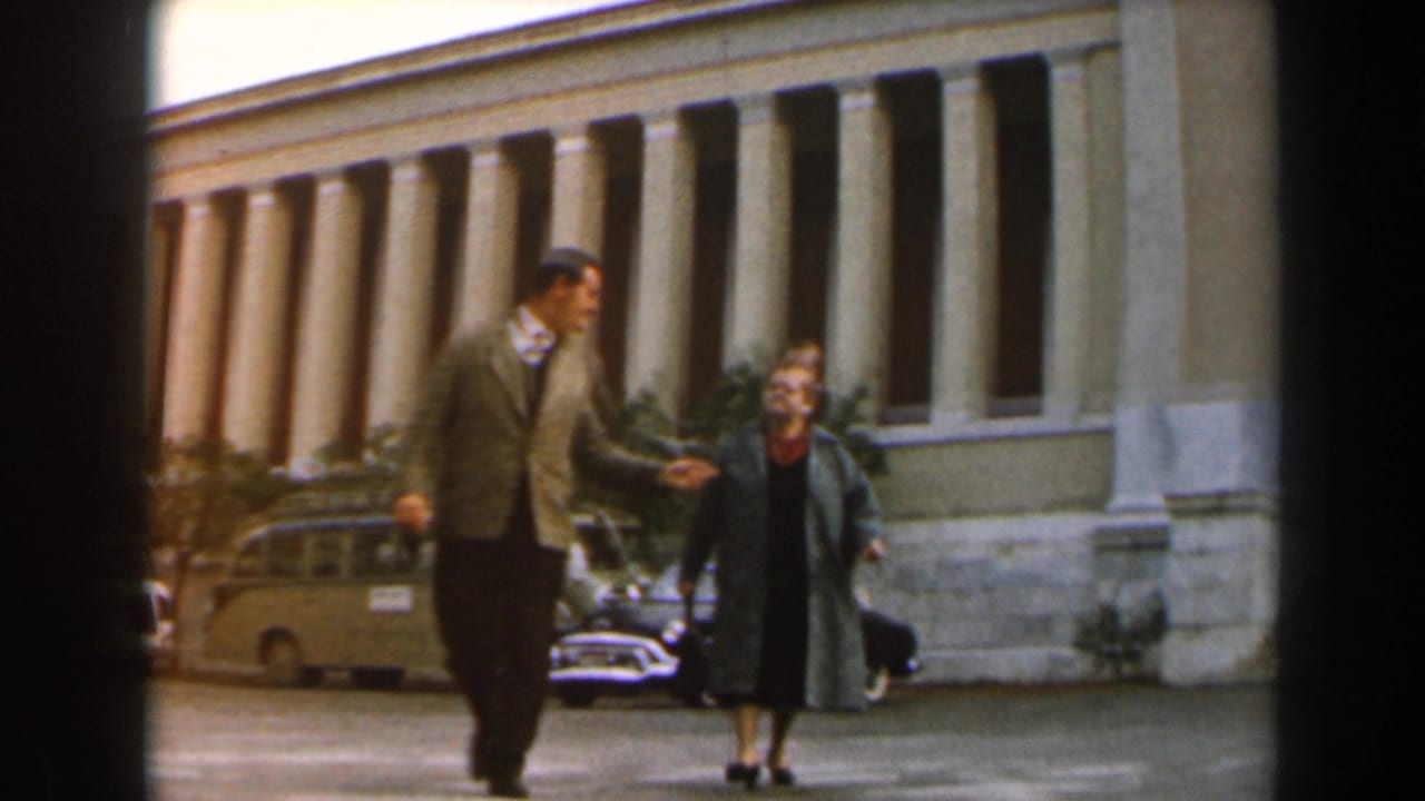 In 1960 Israel footage, an adult man and woman walk past a grand colonnaded building with period cars parked nearby.