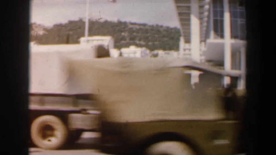 A light-colored period car passes through the foreground, partially obscuring a modernist building and hillside in the background.