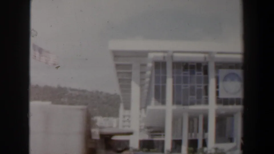 1960 Israel frame of a white institutional-looking facade with tall windows, columns, circular emblem, and hills in the background.
