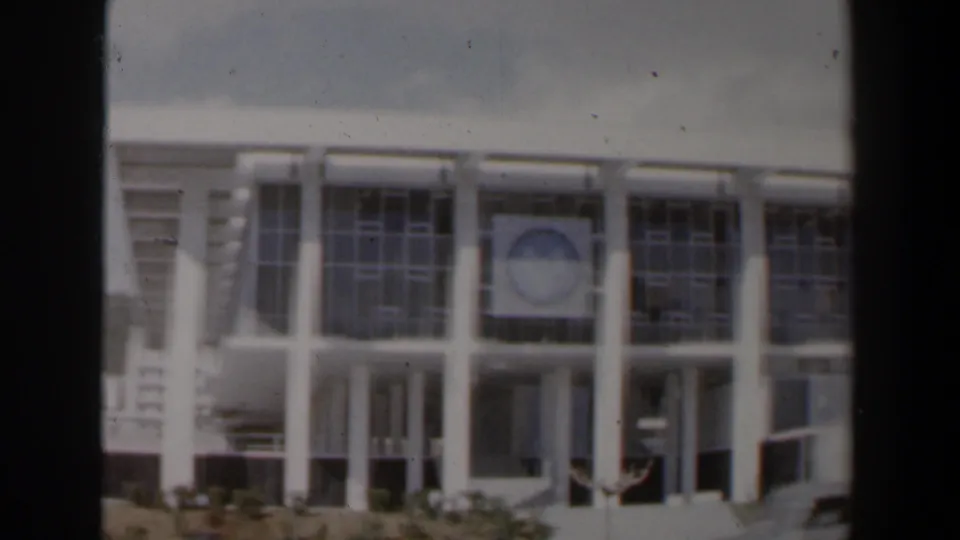 Soft 8mm view of the modernist white facade, showing tall glass panels, columns, and a circular emblem above the entrance area.