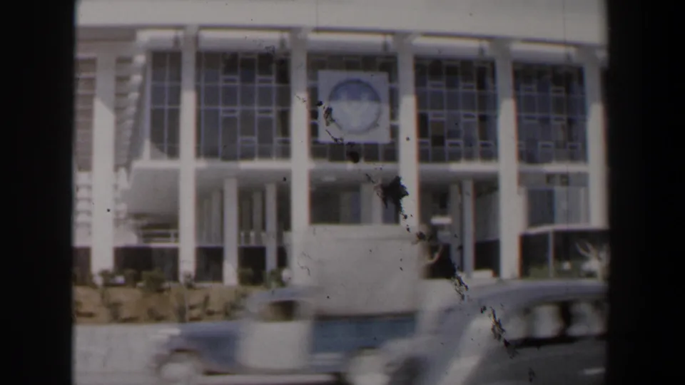 1960 Israel exterior view with the white modernist building, circular emblem, roadside wall, and blurred cars passing in front.