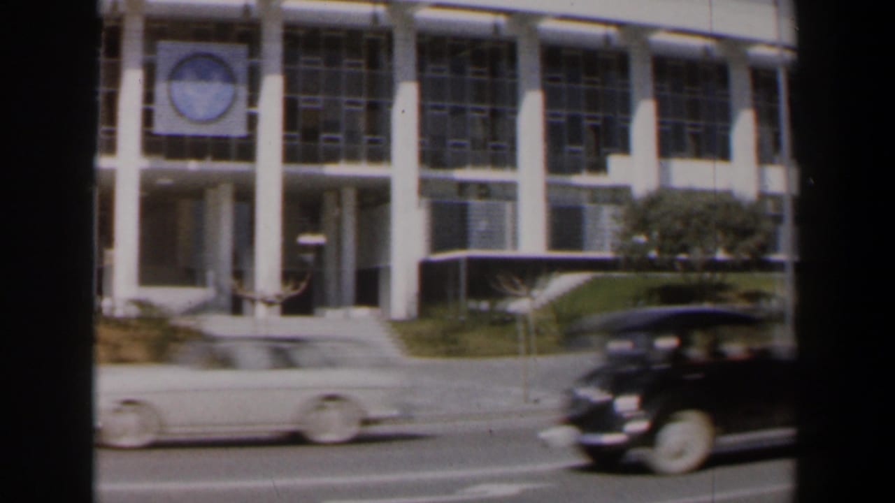 Light-colored car and dark vintage car pass in front of the columned building.
