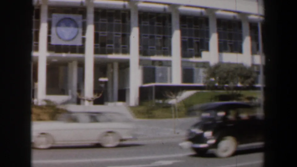 Classic cars move along an Israeli street in front of a modernist building with a visible circular wall emblem.
