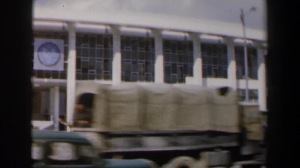 A blurred truck crosses the foreground of the 1960 Israel street scene, obscuring part of the modernist facade.