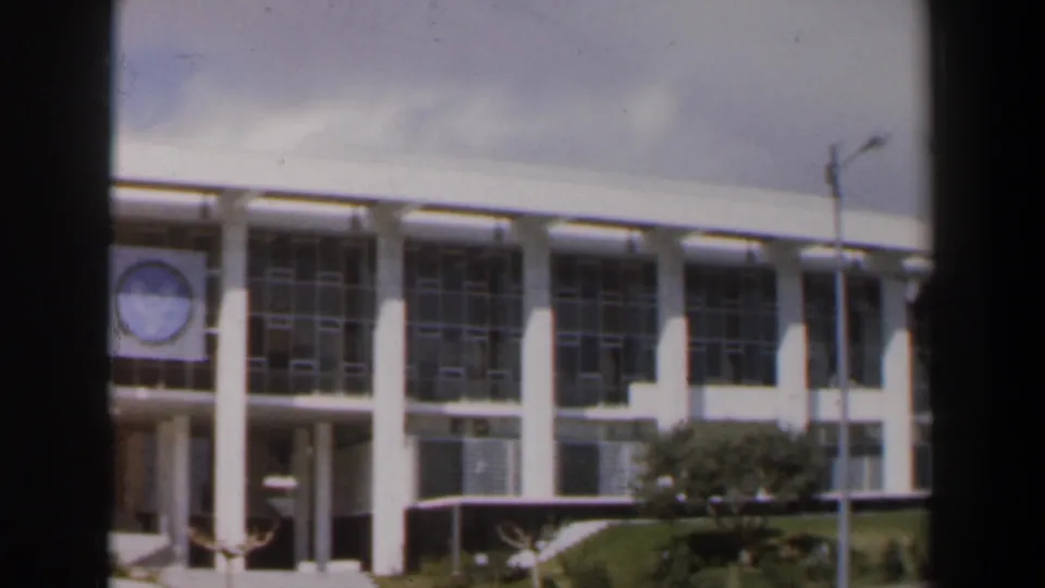 The camera returns to the modernist Israeli building facade with gridded windows, white columns, landscaping, street, and light pole.