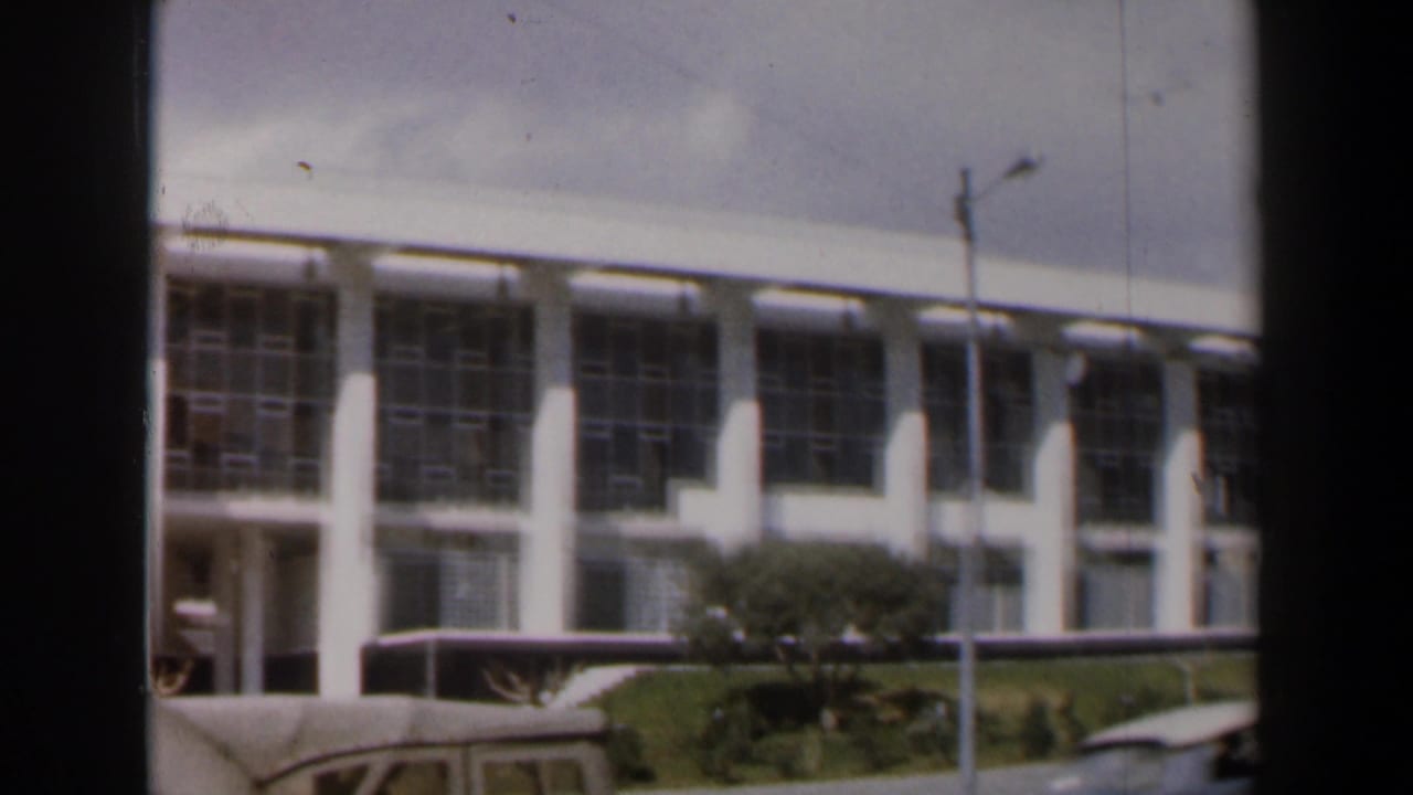 White modernist building with large windows behind a road with a bus roof and car edge.