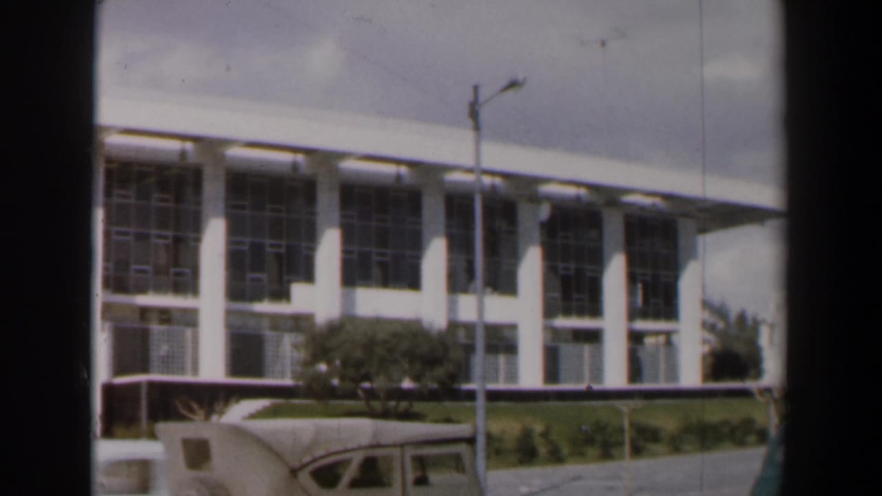 White building facade with columns, windows, streetlamp, shrubs, and partial bus roof.