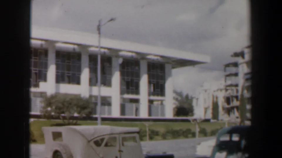 1960 Israel street scene with a white modernist building, streetlamp, military-style jeep, and mid-century cars in traffic.