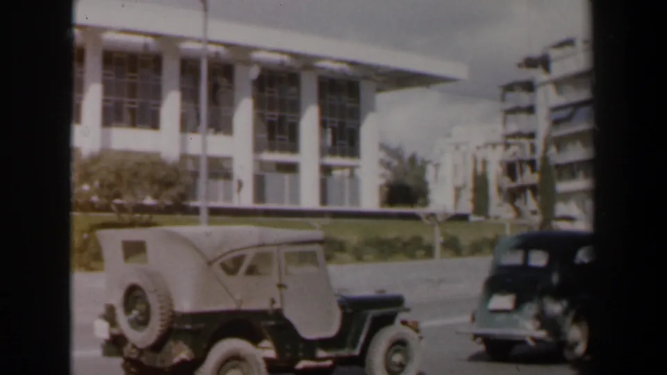 A military-style jeep and dark mid-century car move past a white modernist building in 1960 Israel.