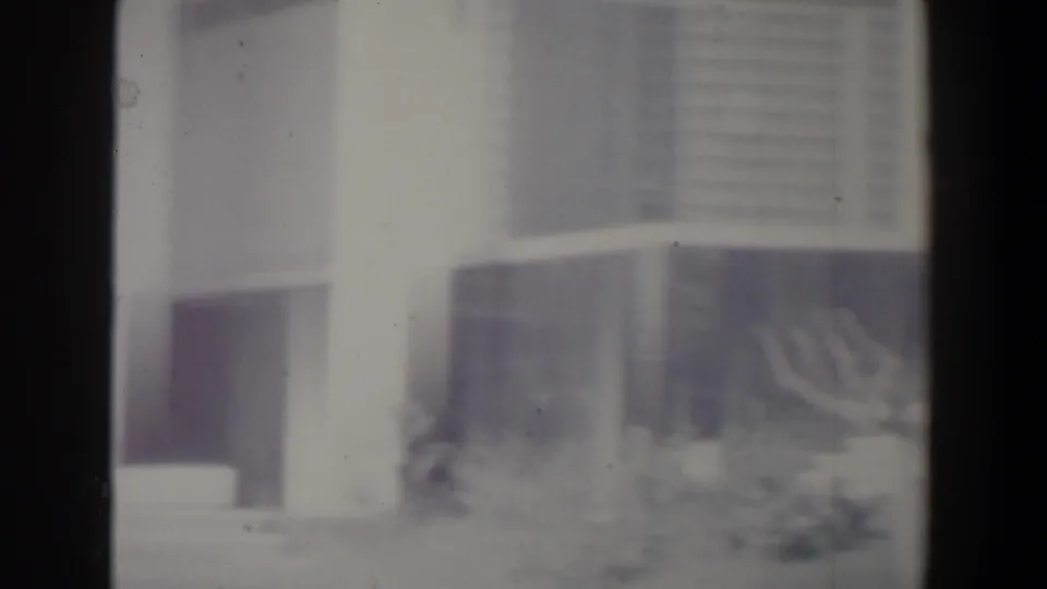 1960 Israel home-movie frame showing a hazy white modern building facade with windows, shrubs and dark film-frame edges.