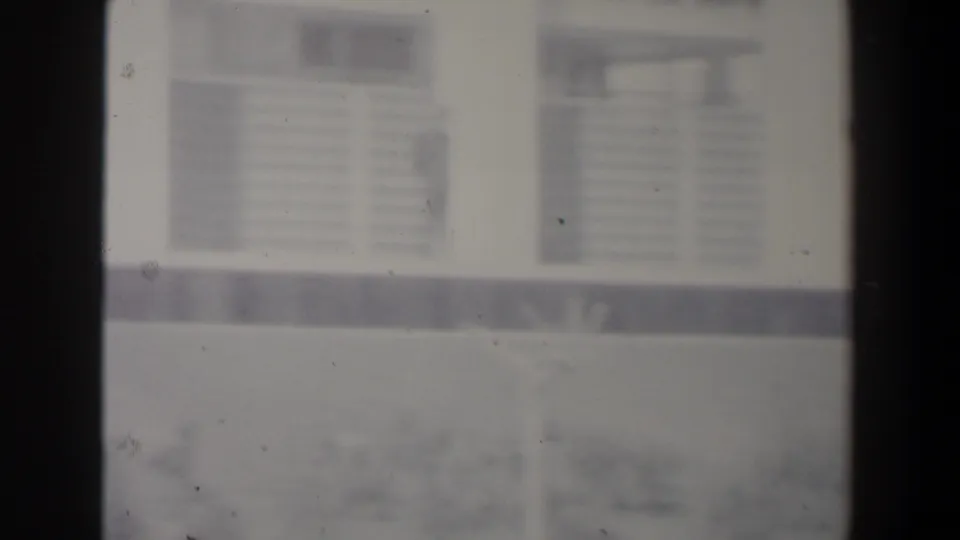 1960 Israel frame showing a hazy white modern building, louvered windows, low wall and faint tree branches in the foreground.