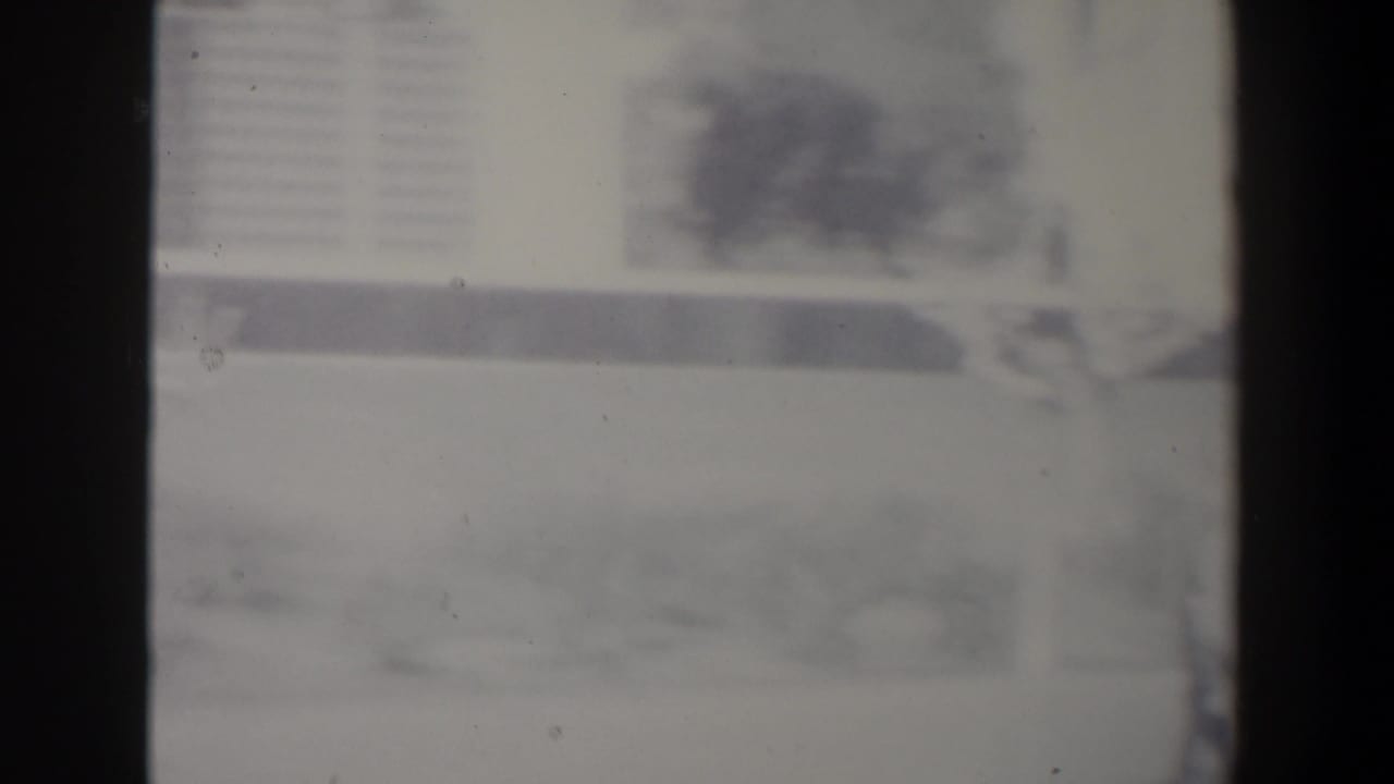 Soft black-and-white 8mm frame from Israel showing an indistinct urban street, vehicle form, buildings, and heavy film grain.