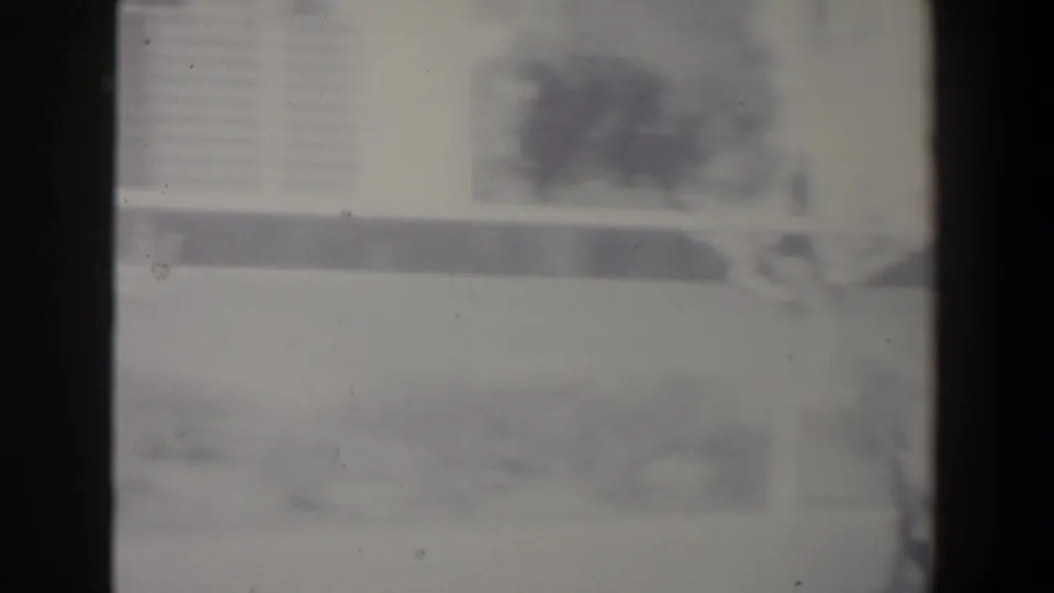 Soft black-and-white 8mm frame from Israel showing an indistinct urban street, vehicle form, buildings, and heavy film grain.