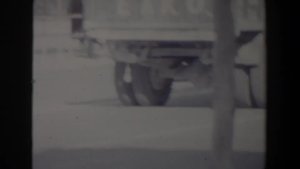 Black-and-white 8mm frame from Israel showing a boxy truck or van, a blurred pedestrian, street surface, and building frontage.