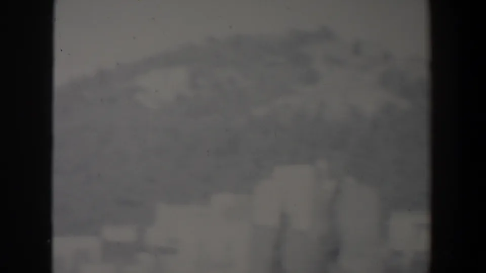 1960s Israel home-movie frame showing blurred hillside buildings below a hazy mountain slope, divided by dark foreground bars.