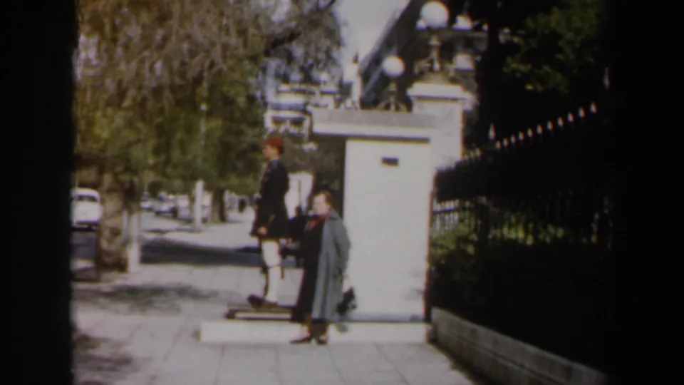 1960 Israel home-movie frame of a guard beside a sentry box, with a woman on the sidewalk and cars beyond the trees.