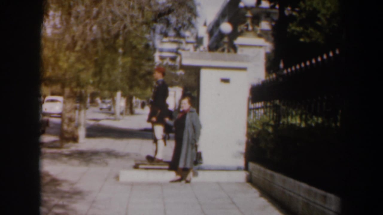 Woman in a long coat walks past a uniformed guard beside a white sentry box.
