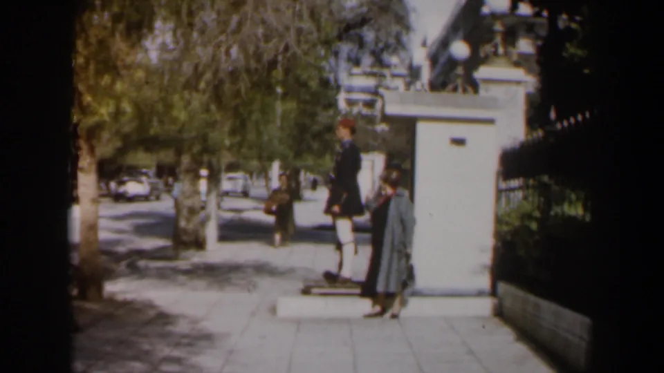 1960 Israel home-movie frame of a sidewalk guard post with a passing woman, distant pedestrian, trees, and older cars.