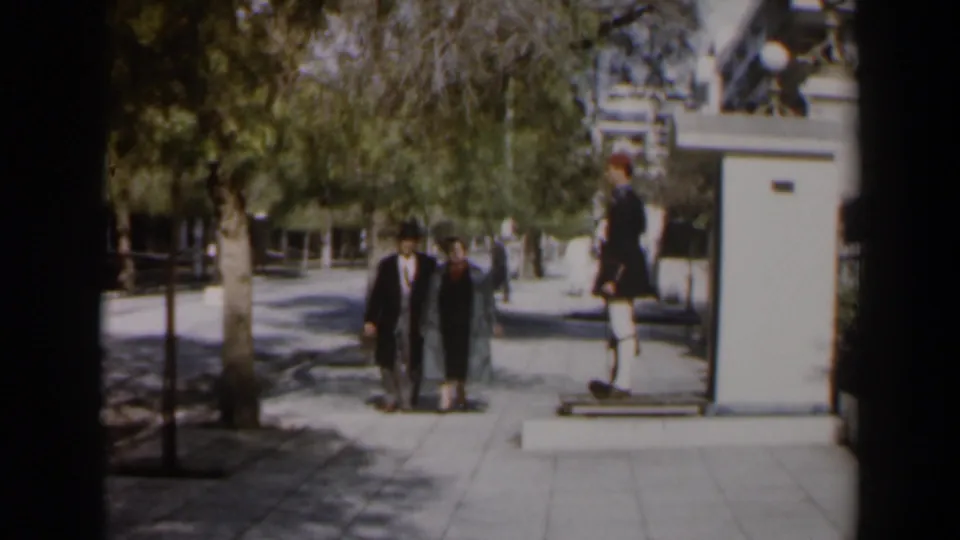 The couple moves closer on a sun-dappled walkway in Israel, with trees overhead and a uniformed guard stationed at the right.
