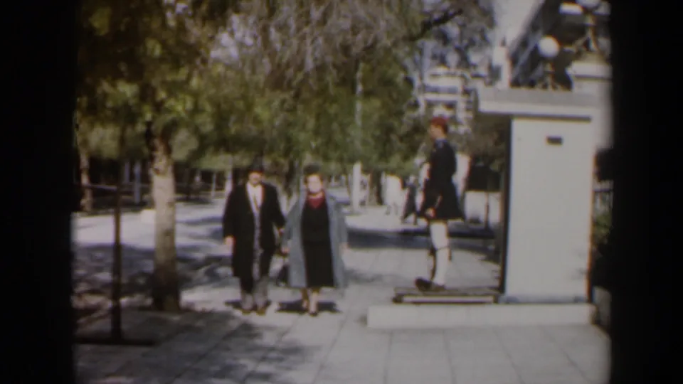A suited man and coated woman walk toward the camera on a public sidewalk, with a uniformed guard still visible beside the small post.