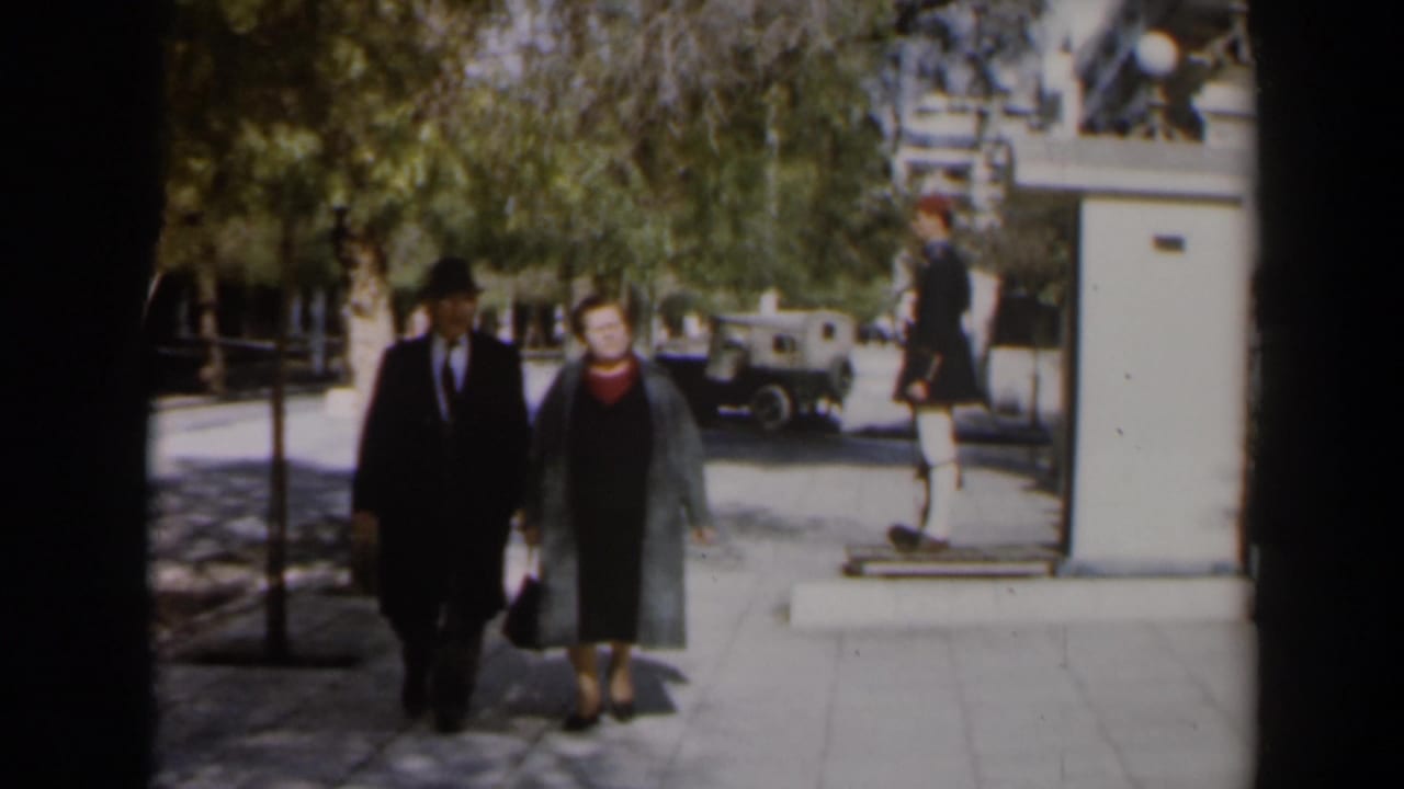 Older couple walking hand-in-hand on a shaded sidewalk near a building entrance.