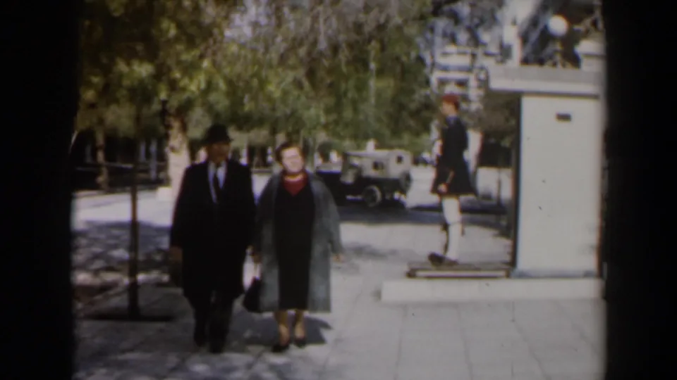 In 1960s Israel, an older couple walks together along a tree-lined sidewalk with a suited man and a woman in a long coat.