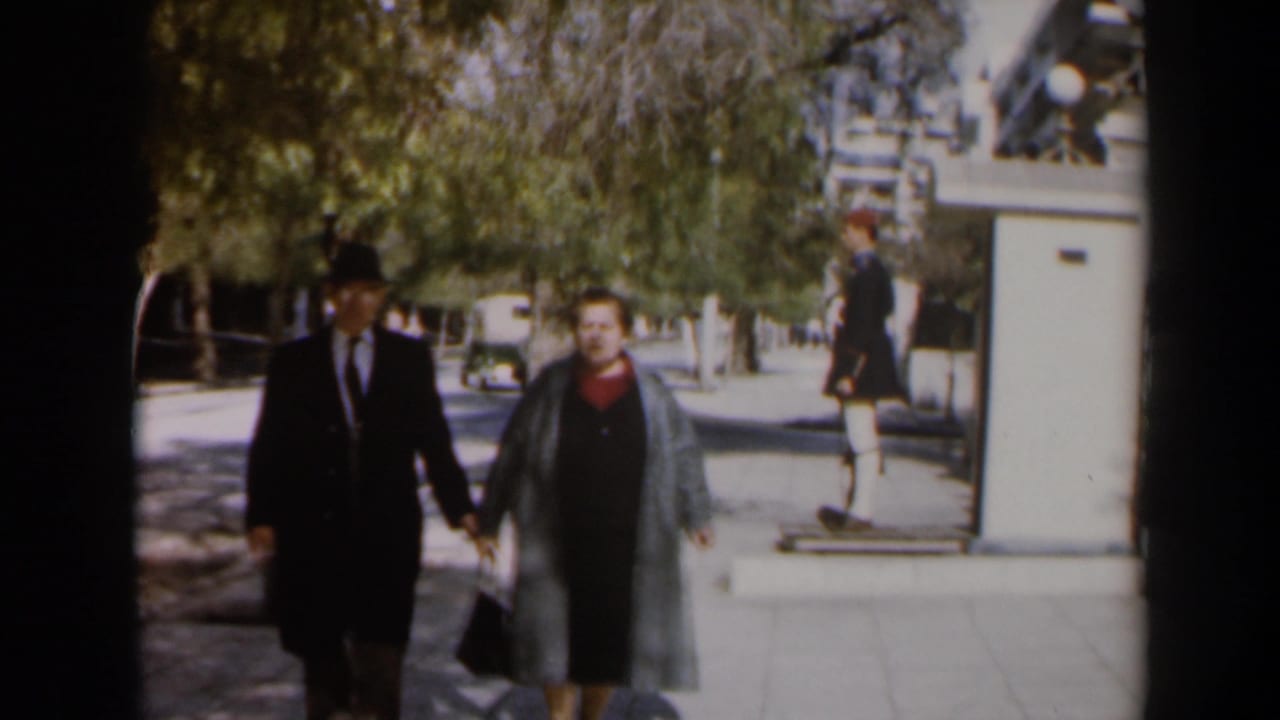 Older couple approaches the camera while a uniformed figure stands near the entrance behind them.