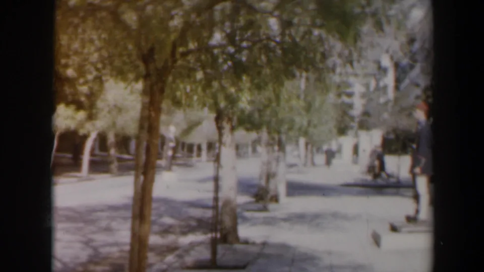 1960 Israel home-movie frame of a leafy street, a vehicle on the road, and a pedestrian near the right edge.