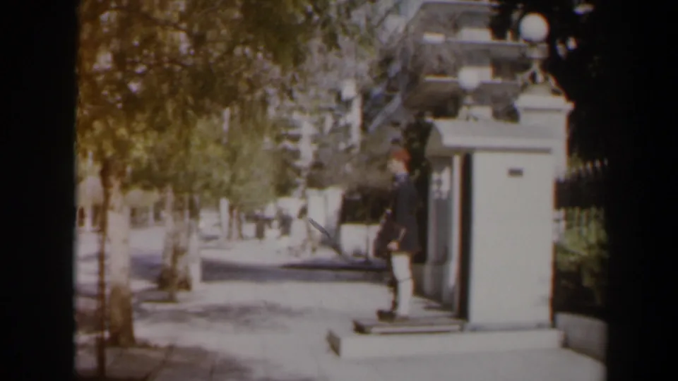 1960 Israel street frame showing a uniformed man standing near a small booth beside trees, sidewalk, and roadway.