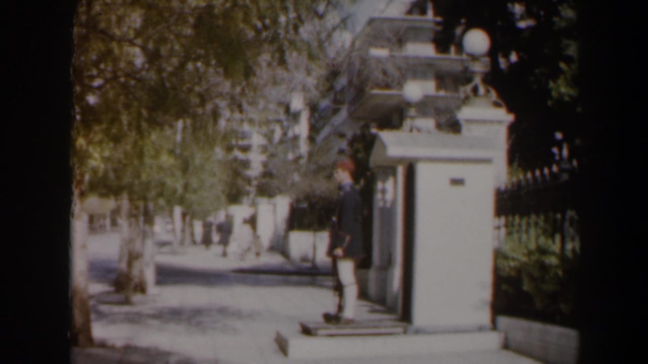 1960 Israel 8mm frame showing a uniformed guard by a white sentry box as pedestrians walk along a shaded sidewalk.
