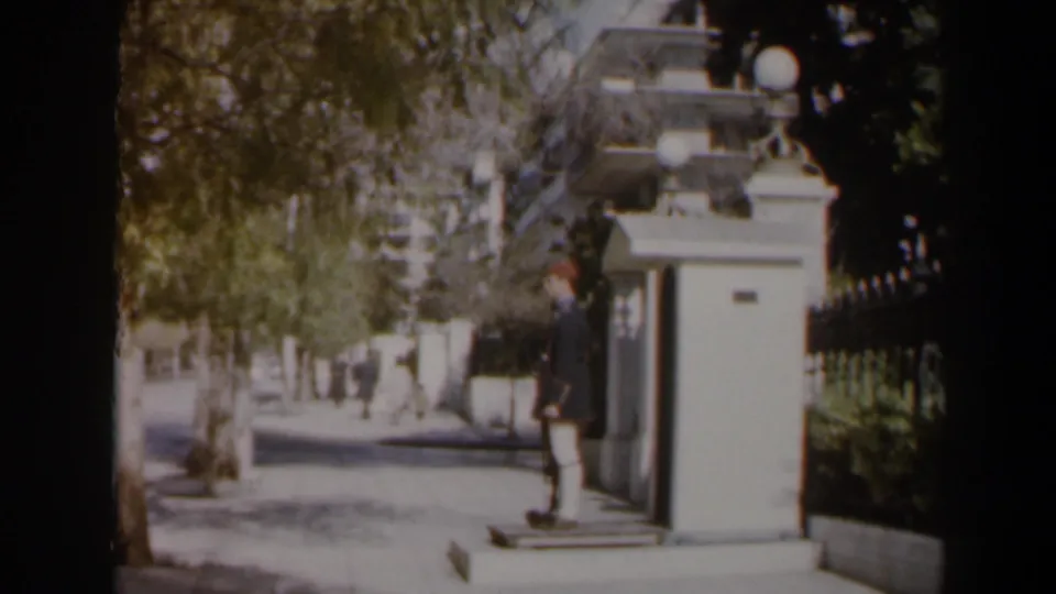 1960 Israel 8mm frame showing a uniformed guard by a white sentry box as pedestrians walk along a shaded sidewalk.