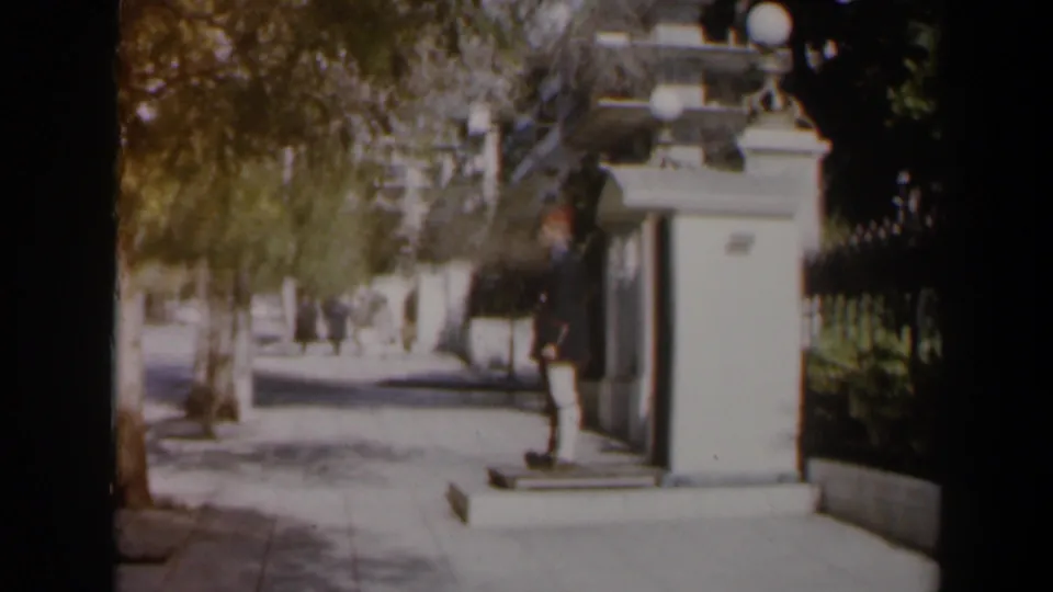 Vintage 8mm frame from Israel showing a guard at a sidewalk sentry box with pedestrians and street trees in view.