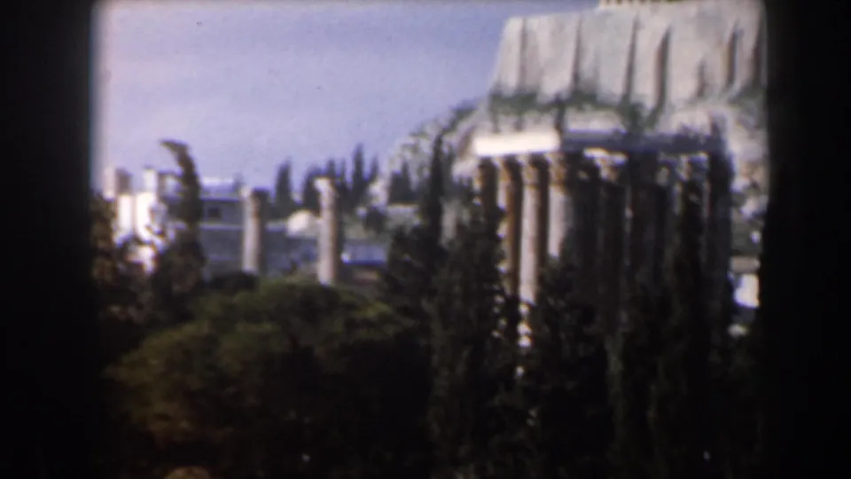 1960s travel footage frame showing classical columns and pale stone ruins partly obscured by tall trees and dark foreground edges.