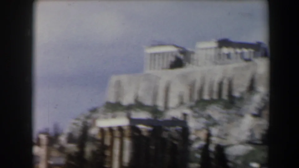 Soft 8mm frame of pale classical ruins on a rocky elevated site, with columns below and open sky behind.